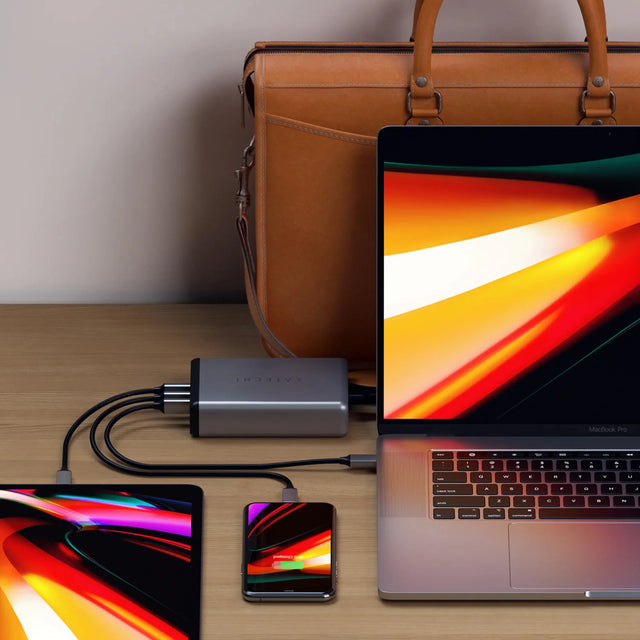 Laptop, tablet, and smartphone connected to Satechi 108W Pro USB-C PD Desktop Charger on a wooden surface with a brown leather bag in the background.