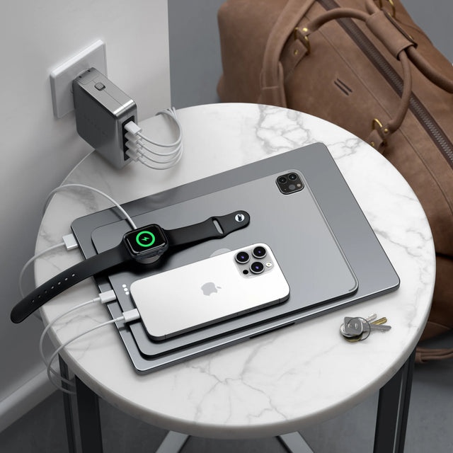Satechi 145W GaN travel charger plugged into a wall outlet charging multiple devices on a small table, including a MacBook, iPad, iPhone and Apple Watch.