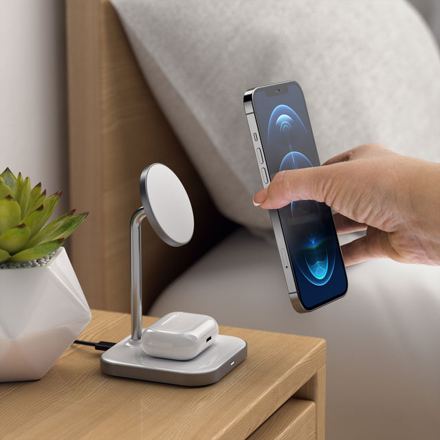 A hand placing an iPhone onto the Satechi Aluminum 2-in-1 Magnetic Wireless Charging Stand while charging  AirPod on a beside table.