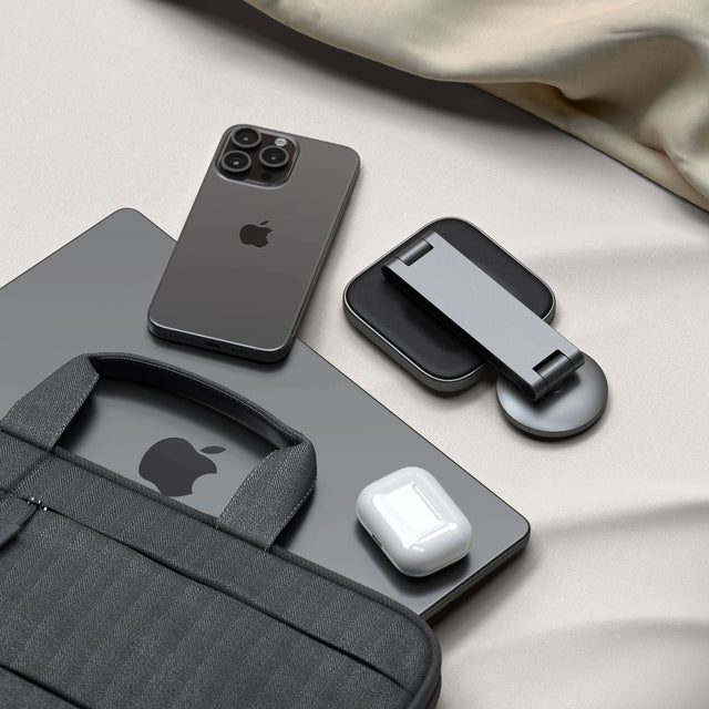Collection of Apple products including an iPhone, AirPods, MacBook, and folded 2-in-1 Foldable Qi2 Wireless Charging Stand on a light surface.