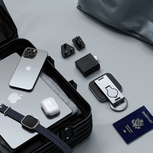 The Satechi 3-in-1 Foldable Qi2 Wireless Charging Stand folded on the bed, next to its travel power adapters and Passport, next to suitcase with iPhone, Apple Watch, and AirPods.