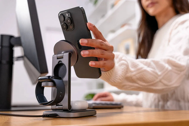 Hand placing an iPhone onto a Satechi 3‑in‑1 Foldable Qi2 Wireless Charging Stand on a desk, showing magnetic phone charging, Apple Watch charging dock, and AirPods charging base.