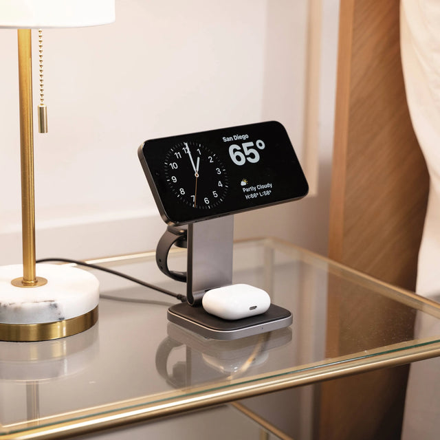 Satechi 3-in-1 Foldable Qi2 Wireless Charging Stand charging an iPhone with digital display and clock, next to a lamp and marble base.