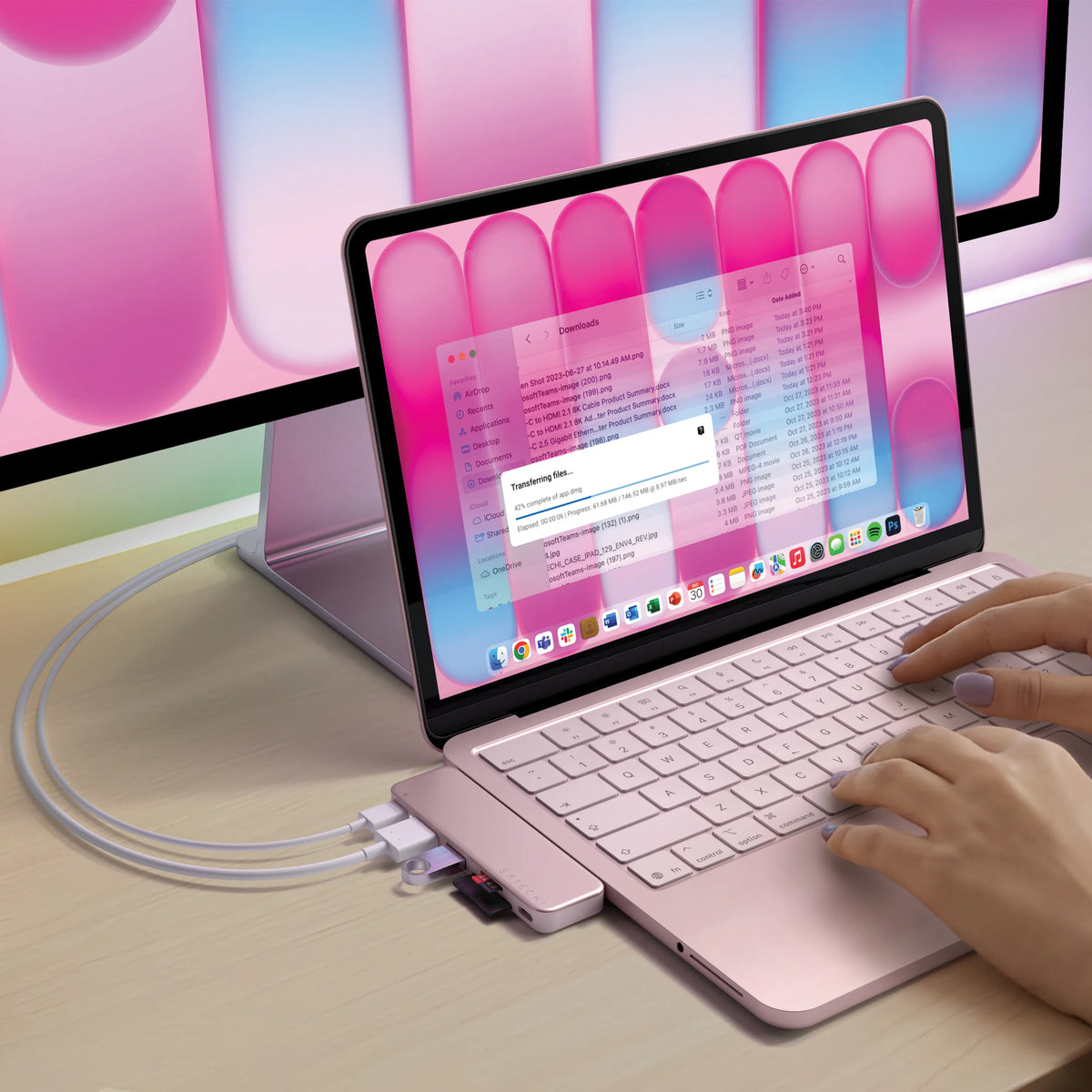 Blush  Satechi USB-C Snap Hub attached to the side of a matching Blush MacBook Neo as a person types, the hub's two USB-C connectors plugged in and cables running to an external display behind it. The laptop screen shows a Finder "Downloads" window with a file-transfer progress dialog reading "Transferring…" at "42% complete."