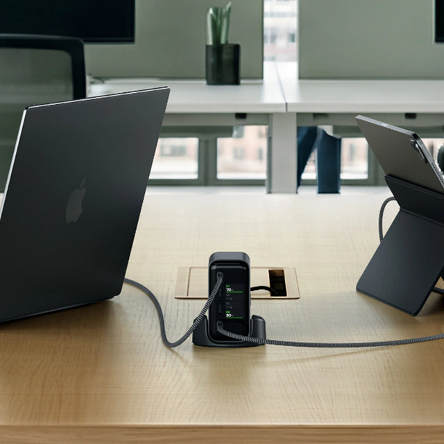 Satechi ChargeView™ 140W Desktop Charger placed at the center of a shared office desk, connected via USB‑C cables to two MacBooks positioned on the left and right. The charger sits upright in a stand with a front‑facing LCD display showing illuminated green indicators, demonstrating desktop charging for multiple devices in a professional workspace.