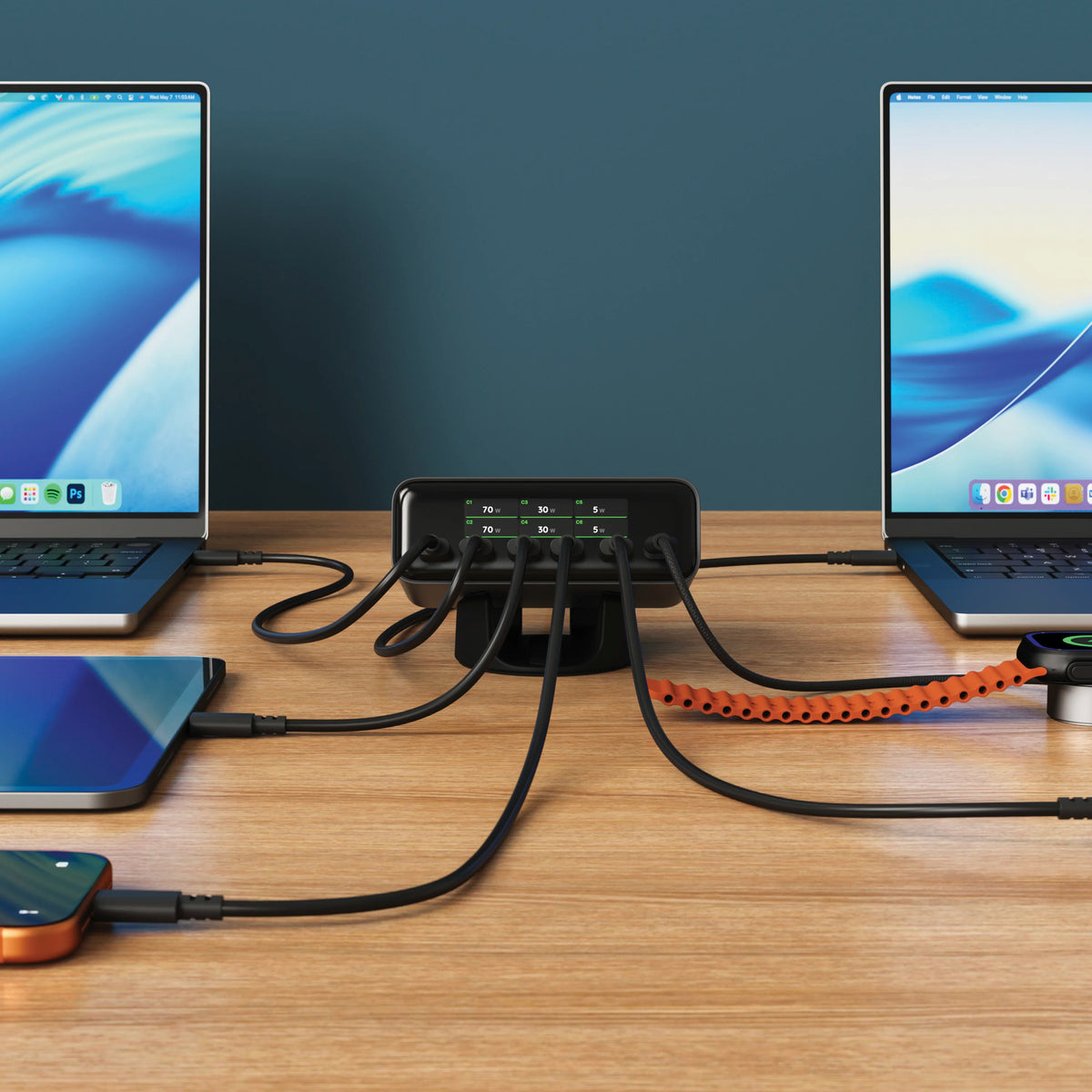 Satechi ChargeView™ 240W Desktop Charger in black positioned horizontally on a wood desk against a dark teal wall, acting as a charging station for multiple devices with six USB-C cables fanning out to a MacBook on the left, a MacBook on the right, a tablet, an orange iPhone, and an Apple Watch charging puck connected by an orange braided cable. The front-facing display of the 6-port USB-C charger reads: C1 70 W; C2 70 W; C3 30 W; C4 30 W; C5 5 W; C6 5 W. The Apple Watch screen shows a green charging bolt