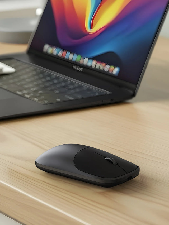 Close-up of the Satechi Slim EX Wireless Mouse in Space Black angled on a light wood surface beside a MacBook displaying a vivid orange and purple wallpaper, with the USB-C charging port visible on the front edge of the mouse. Detail view of the USB-C rechargeable Bluetooth mouse for Mac.