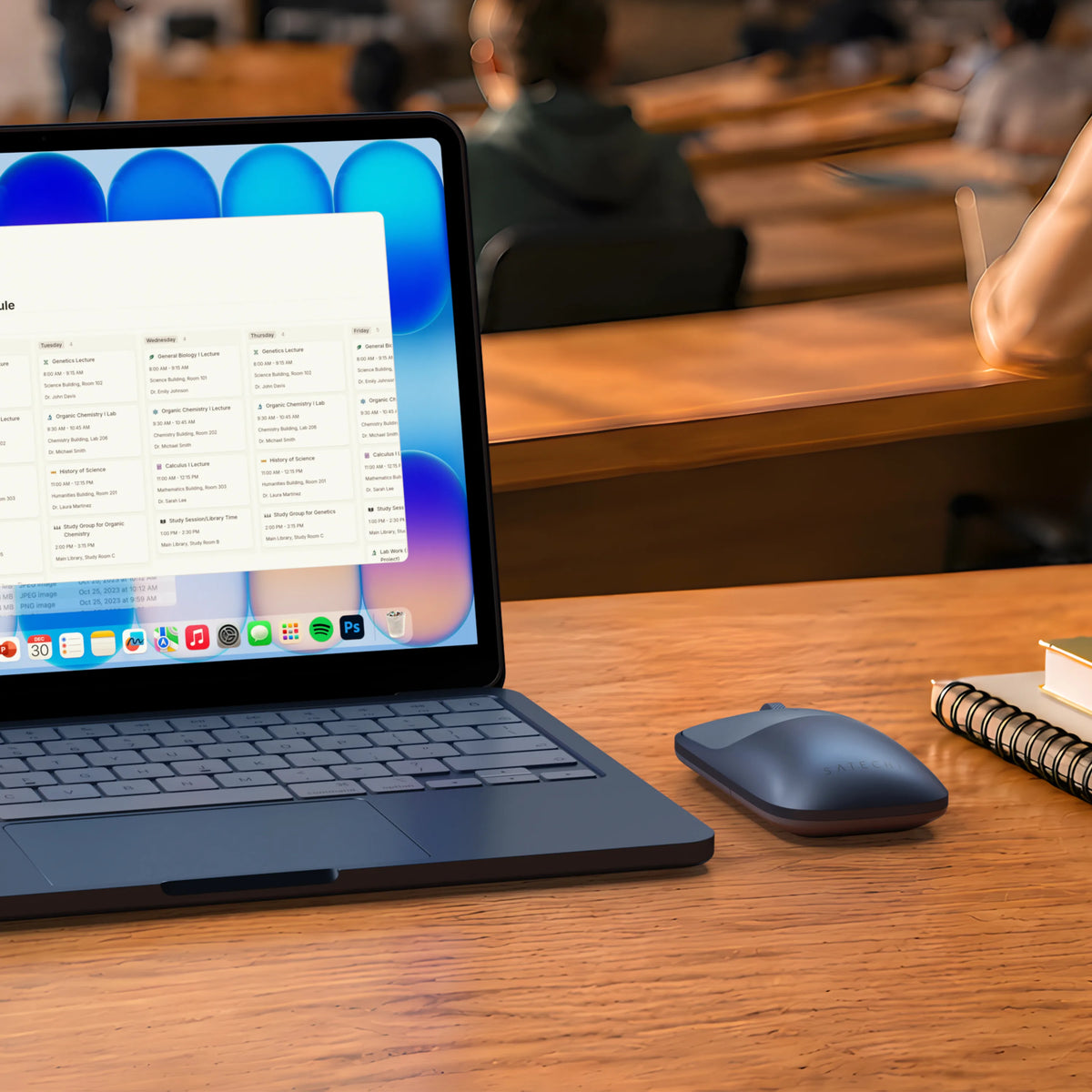 Satechi Slim EX Wireless Mouse in Indigo on a wood desk beside a color-matched Indigo MacBook Neo and a stack of hardcover books in a college lecture hall. 