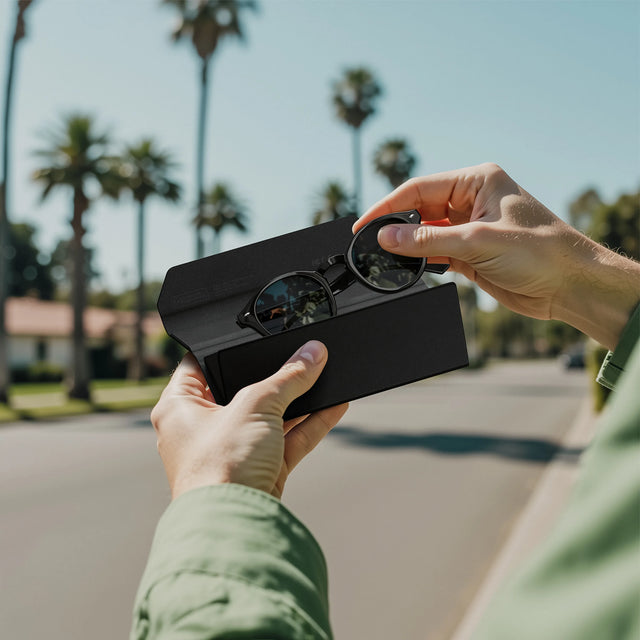 Person sliding their sunglasses into their Vegan-Leather FindAll™ Glasses Case on a street with palm trees in the background.