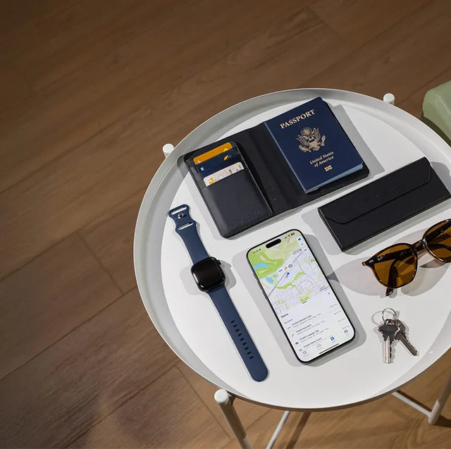 Table with travel essentials including a Vegan-Leather FindAll™ Passport Cover Satechi Black, iPhone, Vegan-Leather FindAll™ Glasses Case,Vegan-Leather FindAll™ Luggage Tag, Apple Watch, keys, and sunglasses on a wooden floor.
