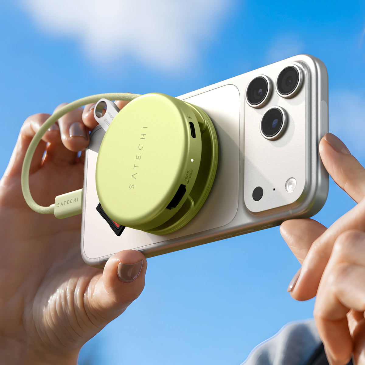 Hands holding a white iPhone Pro against a blue sky with a citrus OntheGo 5-in-1 Multiport Adapter magnetically attached to the back, transferring data from the iPhone to an SD card and a flash drive.