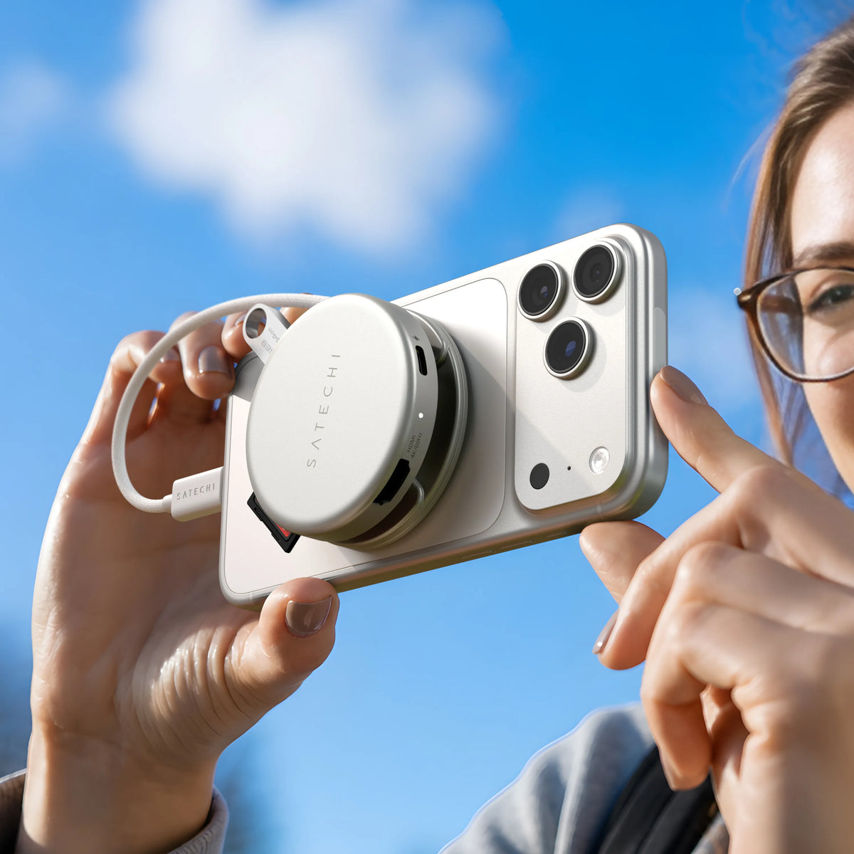 A person's hands hold a white iPhone against a bright blue sky, with a Silver Satechi OntheGo™ 5-in-1 Multiport Adapter attached to the back via MagSafe, its coiled braided cable and a USB flash drive visible. 