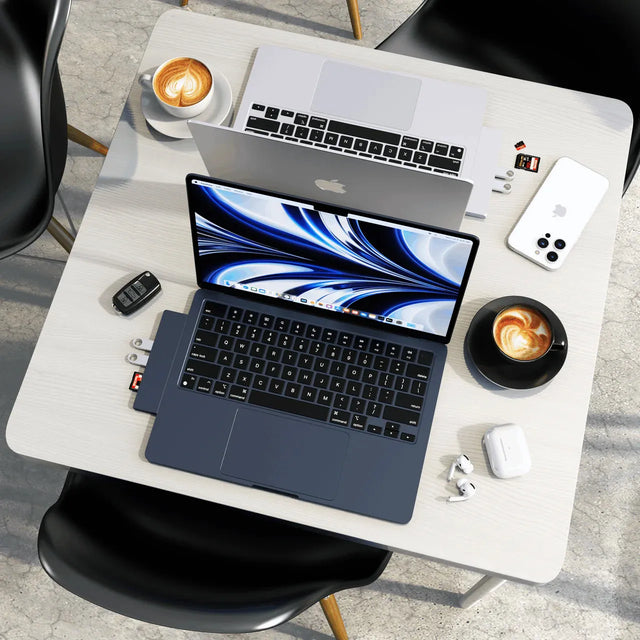 Top‑down view of a workspace with two MacBook Air laptops, both using the Pro Hub Slim attached and uploading files from flash drives and SD cards , surrounded by accessories including an iPhone, AirPods, and coffee cups.