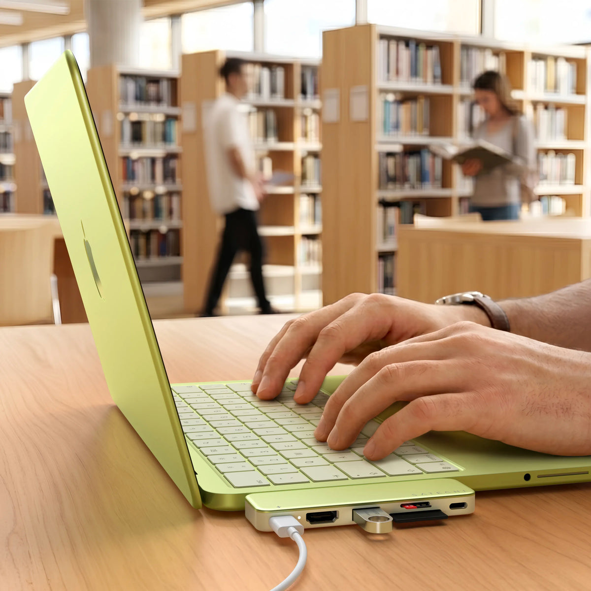 Satechi USB-C Snap Hub in Citrus green attached to the side of a color-matched Citrus MacBook Neo on a light wood library table, with a person's hands typing on the keyboard. The hub's ports are in use and visible along the front edge — a white USB-C cable plugged in on the left, an HDMI port, a USB-A port with a flash drive inserted, an SD and microSD card slot with red accents, and a USB-C port — with the "SATECHI" wordmark printed along the top of the aluminum housing. 