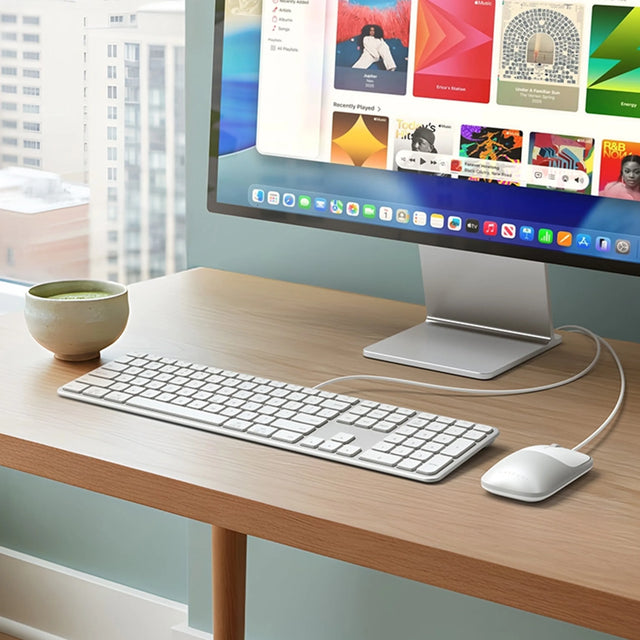 Slim LX3 Wired Keyboard and Slim LX Wired Mouse displayed on a modern wood desk setup with a widescreen monitor, showing a clean and minimalist workspace ideal for productivity. The ultra‑slim wired keyboard and mouse are positioned neatly beside a ceramic coffee cup and stacked books, highlighting a contemporary home office featuring natural light and desktop accessories.