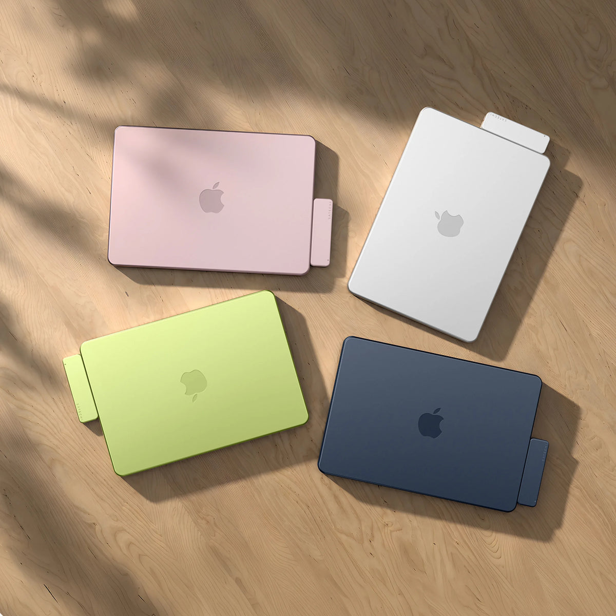Four MacBooks — Blush, Silver, Citrus , and Indigo — arranged top-down on a sunlit wood table, each with a color-matched Satechi USB-C Snap Hub attached to its side edge. 