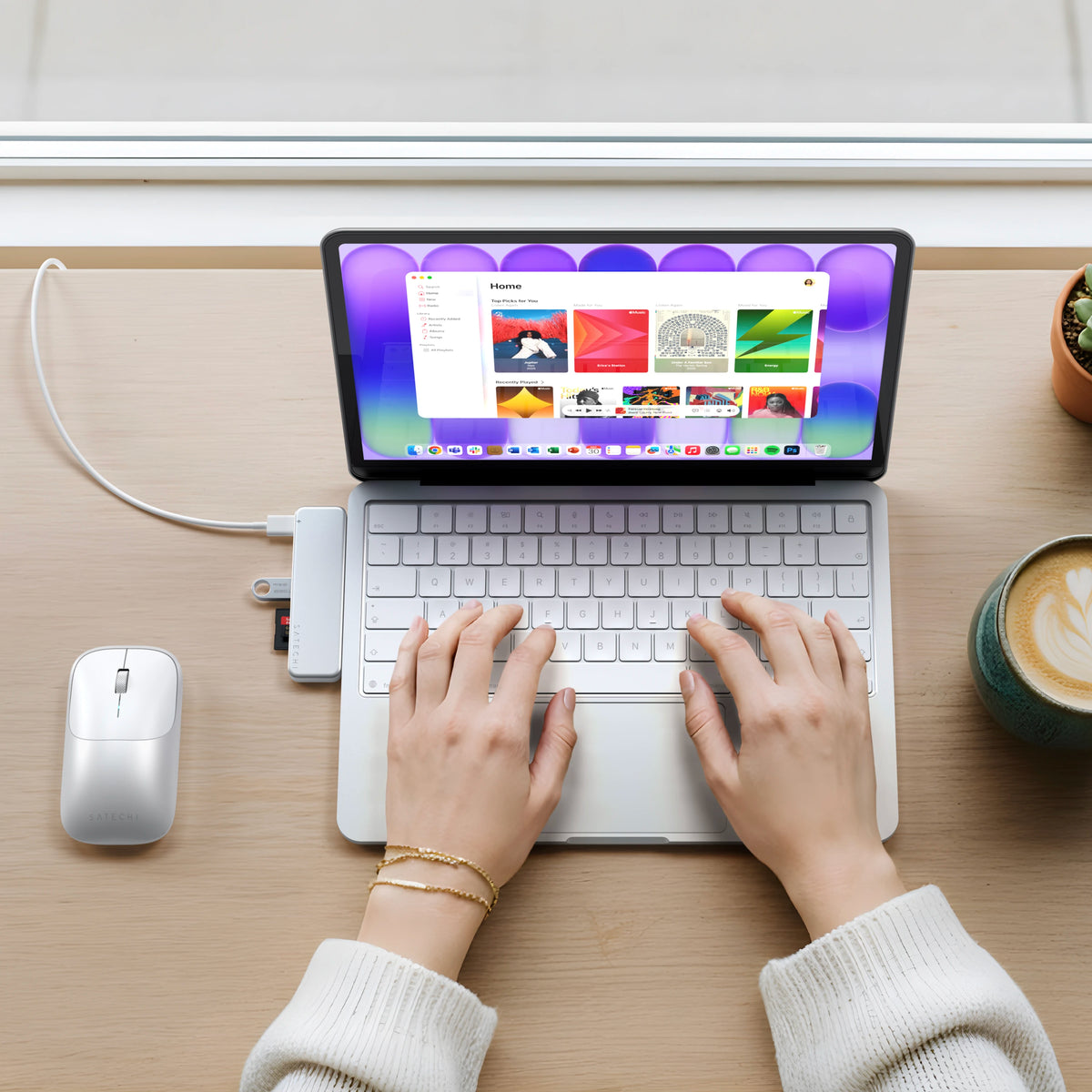 Hands with a gold bracelet typing on a silver MacBook Neo, with a silver USB-C Snap Hub connected to transfer files and deliver pass-through charging, a silver Slim EX wireless mouse for Mac alongside, and a small plant on the wood desk.
