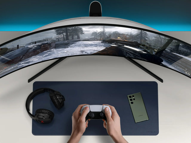 Vegan‑Leather Premium Desk Mat in navy set beneath a curved monitor, gaming controller, headphones, and smartphone for a clean gaming workspace.