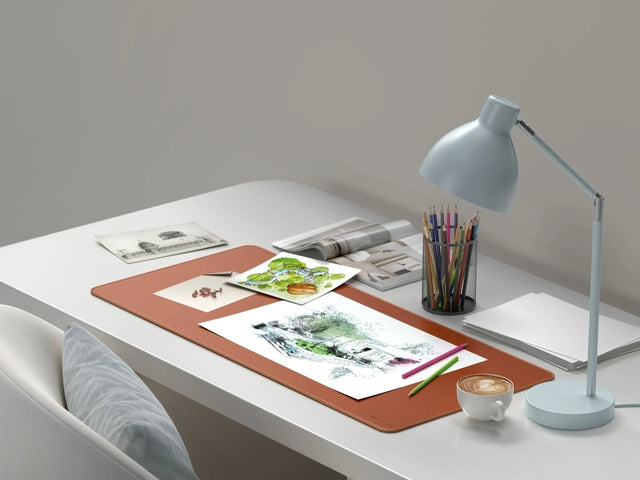 Vegan‑Leather Premium Desk Mat in brown on a white desk, supporting sketchbooks, colored pencils, and artwork beneath a task lamp and coffee cup.