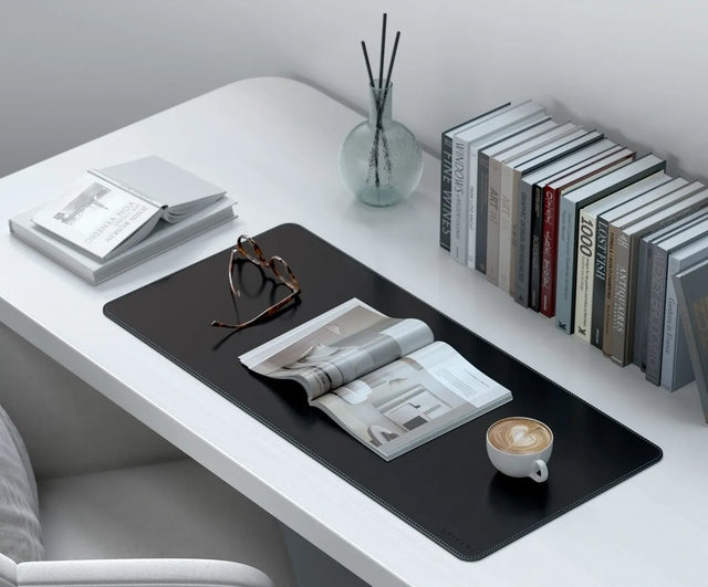 Vegan‑Leather Premium Desk Mat used as a surface for reading materials, glasses, and a cup of coffee on a minimalist white desk.