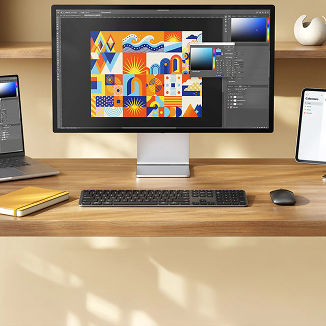 Minimalist workspace with the Slim EX3 Wireless Keyboard centered on a wooden desk and the Slim EX Wireless Mouse placed to the right. A laptop displaying colorful graphics sits to the left, while a large external monitor and a tablet stand are positioned in the background. A yellow notebook rests near the keyboard, creating a clean, modern productivity setup.