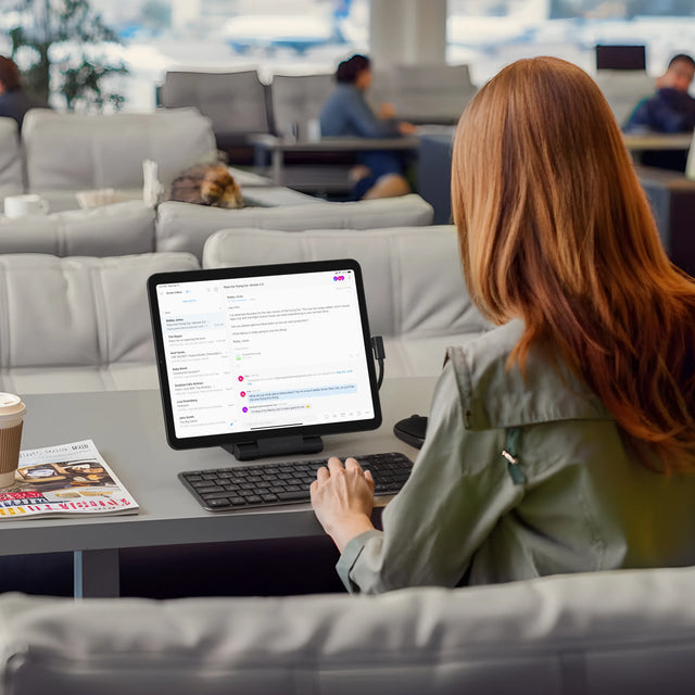 An iPad mounted on the Satechi OntheGo™ Foldable Stand Hub on a lounge-style table, paired with a wireless keyboard. A coffee cup and magazine are placed nearby, showing a portable workstation setup ideal for travel or remote work.