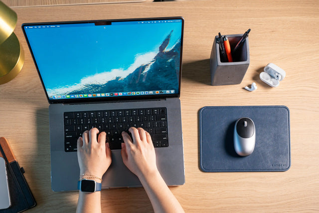 Vegan‑Leather Premium Mouse Pad placed beside a MacBook on a wooden desk, with a wireless mouse resting on the pad and hands typing on the keyboard.