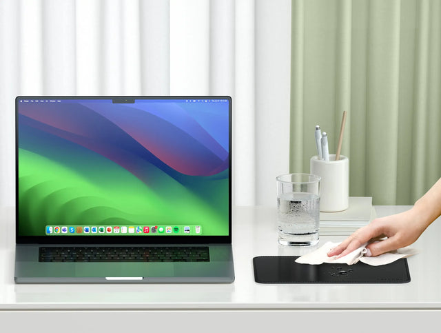 Vegan‑Leather Premium Mouse Pad on a clean white desk as a hand wipes its water‑resistant surface, shown next to a laptop, glass of water, and pen holder.