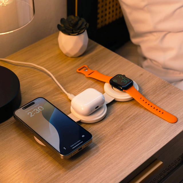 An iPhone, Apple Watch, and AirPods on a OntheGo™ 3-in-1 Chargers, charging simultaneously while on a bedside table. 
