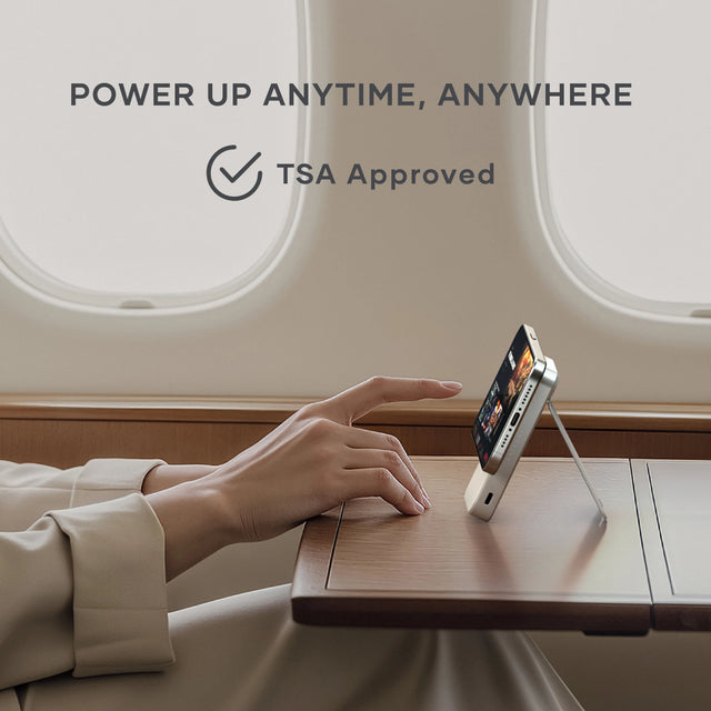 Satechi Onthego Power Bank 10K supporting a smartphone upright on an airplane tray table, emphasizing TSA-approved portability for travel. Text says: Power Up Anytime, Anywhere. TSA Approved.