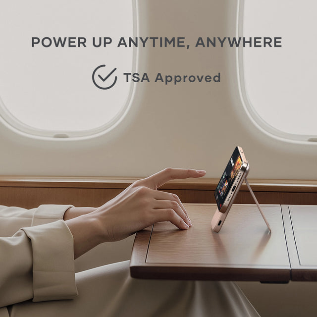 Satechi On-the-Go 5K Power Bank supporting a smartphone upright on an airplane tray table, emphasizing TSA-approved portability for travel. Text says: Power Up Anytime, Anywhere. TSA Approved.