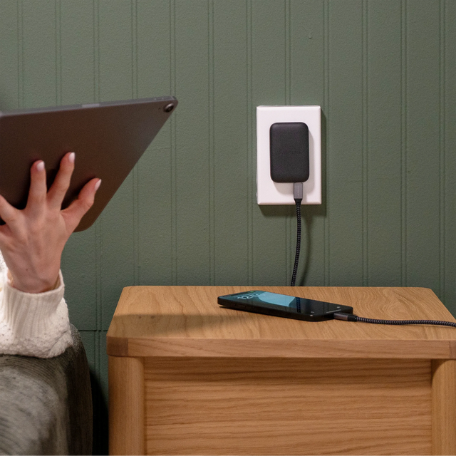 Person using a tablet while their iPhone charges with a OntheGo™ 67W Slim Wall Charger against a green wall.