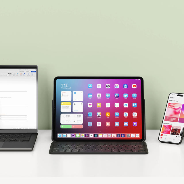 An iPad on the OntheGo™ Bluetooth Keyboard with Stand, next to a MacBook, and iPhone on a white surface with a light green wall back.