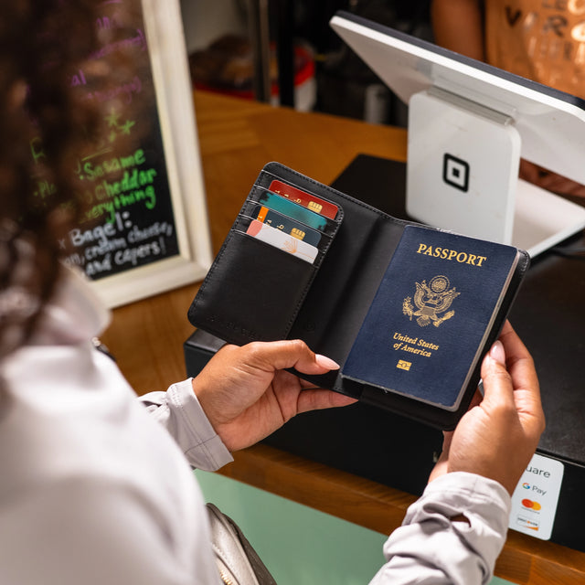 Person holding a Vegan-Leather FindAll™ Passport Cover with passport and cards inside, near a counter with register.