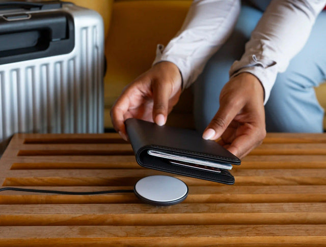 Person aligning a Vegan‑Leather FindAll™ Passport Cover onto a magnetic charging pad on a wooden surface, with a suitcase blurred in the background.