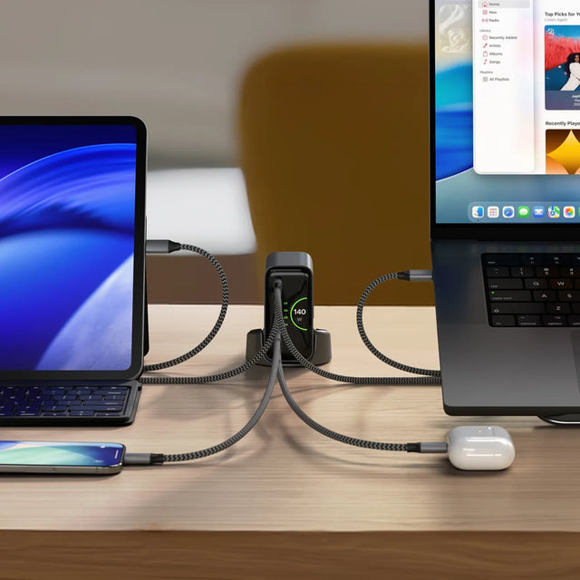 Satechi ChargeView™ 140W Desktop Charger positioned at the center of a desk, powering two MacBooks and AirPods simultaneously via braided USB‑C cables. The charger’s front-facing LCD screen displays ‘140’ in green text, indicating real‑time power output while multiple devices are connected.