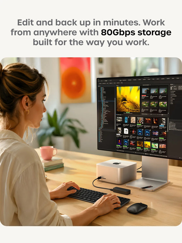 Woman editing photos at a Mac Studio workstation with a large display showing Adobe Bridge filled with RAW image thumbnails, the silver Satechi DotDisk M.2 NVMe enclosure connected nearby. Edit and back up in minutes. Work from anywhere with 80Gbps storage built for the way you work. Silver DotDisk external SSD enclosure shown at an angle with USB-C port visible.