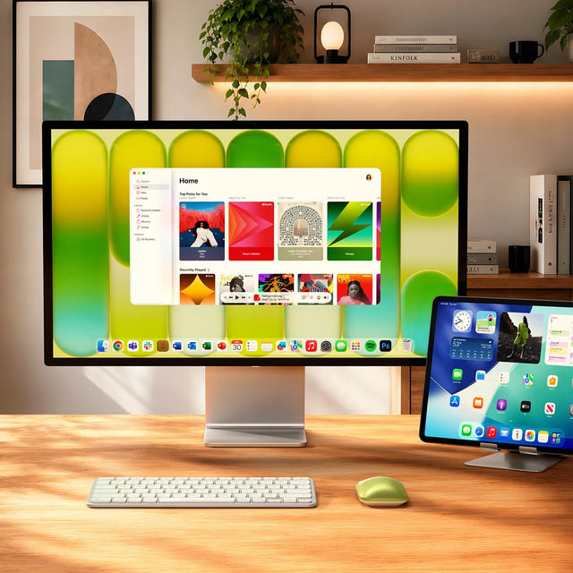 Satechi Slim EX Wireless Mouse in Citrus green on a light wood desk beside a silver Slim EX1 Wireless keyboard, positioned in front of an Apple Studio Display and an iPad on a stand. The display shows the Apple Music app open to "Home" with "Top Picks for You," "Listen Again," "Made for You," "Mood for You," and "Recently Played" sections; a floating shelf behind holds trailing plants, a lamp, a mug, and books including one spine reading "KINFOLK.