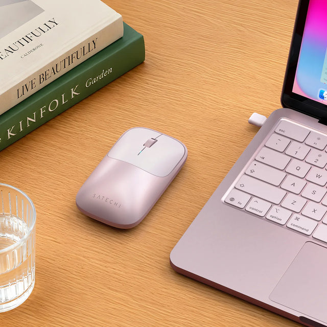 Satechi Slim EX Wireless Mouse in Blush on a wood desk beside a Blush MacBook Neo. The  logo "SATECHI"  shown on the front of the mouse. A glass of water sits to the left and two books rest behind it. The slim wireless mouse styled as a color-matched MacBook Neo accessory.