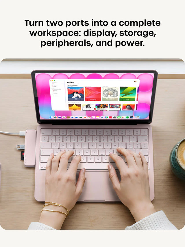 Left: hands typing on a Blush pink MacBook with the Apple Music app on screen and a matching Blush USB-C Snap Hub attached, a USB flash drive and red microSD card inserted. Right: close-up of a Citrus green MacBook with a matching green usb c hub with hdmi, two white USB-C cables, a USB flash drive, and an SD card connected. Headline reads "Turn two ports into a complete workspace: display, storage, peripherals, and power."