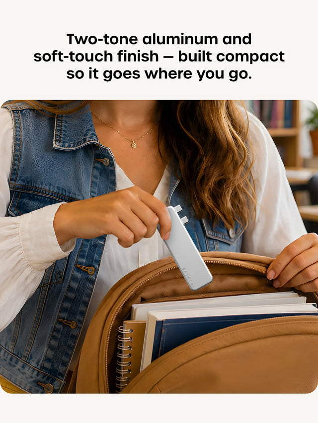 A woman in a denim vest slides a Silver Satechi USB-C Snap Hub into a brown leather backpack packed with notebooks, the SATECHI logo visible on the hub's soft-touch face. Headline reads "Two-tone aluminum and soft-touch finish — built compact so it goes where you go."