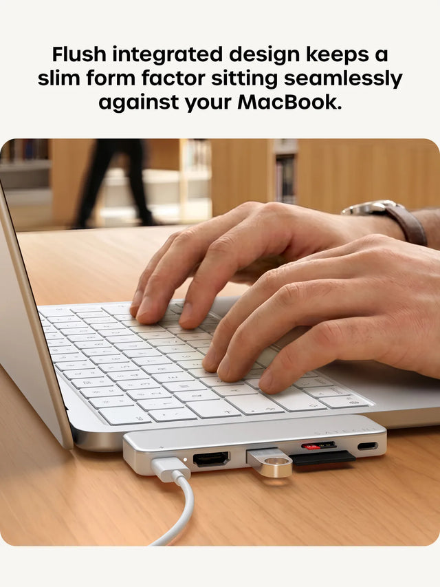 Hands typing on a Silver MacBook in a library with the color-matched Satechi USB-C Snap Hub for MacBook Neo attached to the side. Headline reads "Flush integrated design keeps a slim form factor sitting seamlessly against your MacBook."