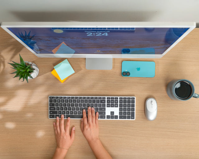 Person using a computer on a wooden desk with a Satechi Slim X3 Bluetooth Backlit Keyboard alongside an M1 Mouse.