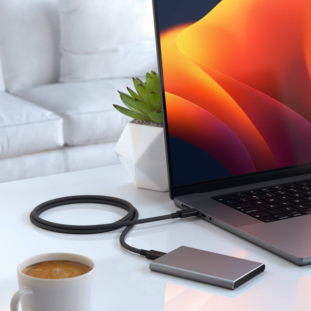 An external hard drive transfering filed via a Thunderbolt 4 Pro Cable - 1M to. MacBook and coffee on a table.