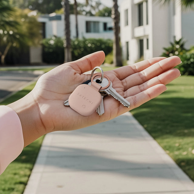 Hand holding a set of keys with a Desert Rose Satechi Vegan‑Leather FindAll Keychain, shown outdoors in front of modern apartments.